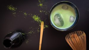 Matcha latte con hielo en una taza azul de cerámica, acompañado de una cuchara y un batidor de de bambú y matcha espolvoreada al lado, sobre un fondo negro