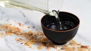 Jarra con caldo dashi japonés siendo vertido en un cuenco negro, con virutas de katsuobushi (bonito seco) al lado, sobre fondo de mármol
