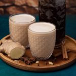 Dos vasos de Chai Latte, una botella del concentrado de chai que compartimos en esta receta, especias y un trozo de jengibre. Receta de chai latte como el de Starbucks