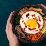 Dos manos sostienen un cuenco de bimbimbap con verduras de colores, ternera en rodajas y un huevo frito cubierto de salsa, visto desde arriba sobre un fondo verde azulado oscuro.