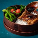 Salsa teriyaki siendo vertida sobre el pollo en un bento japonés con arroz, edamames, ensalada y pollo glaseado con salsa teriyaki - receta casera de salsa teriyaki con pollo glaseado.