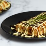 Takoyaki preparado sin sarten especial servido en un plato negro con aderezo de mayonesa y salsa takoyaki, espolvoreado con aonori,