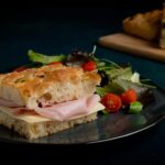 Un sándwich de focaccia relleno de jamón y queso, presentado en un plato negro junto a una ensalada de hojas verdes y tomates cherry.